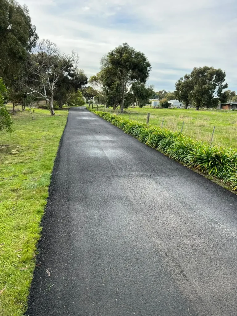 A long, freshly paved asphalt driveway runs through a green rural paddock
